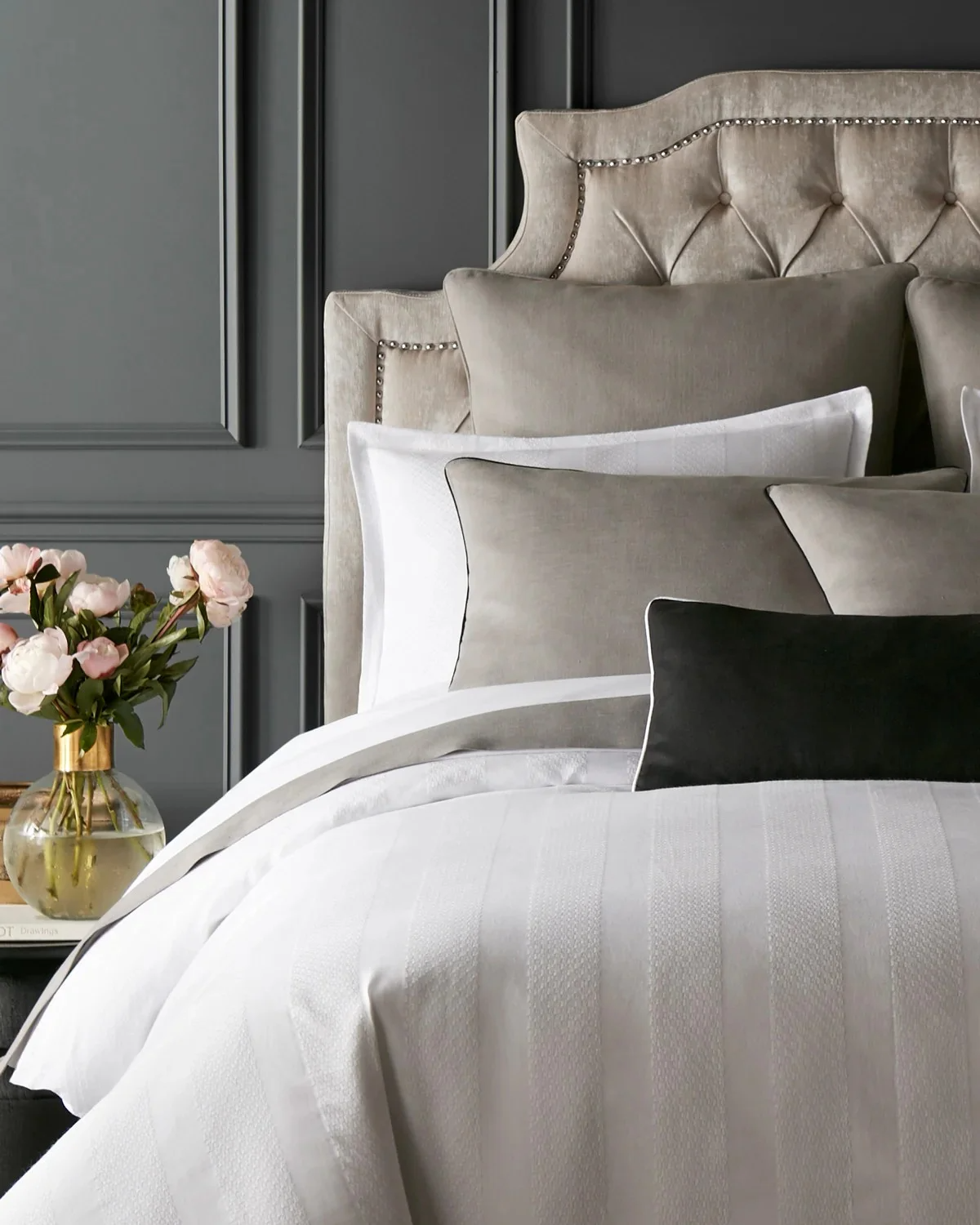 A luxurious bedroom with a tufted beige headboard, accented with decorative metal studs, and a collection of gray, white, and black pillows on a white bedspread. A side table with a glass vase filled with pink peonies is visible.