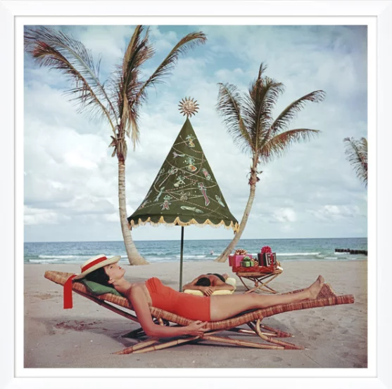 A woman in a red swimsuit and a wide-brimmed hat relaxing on a lounge chair on a sandy beach under a decorated umbrella with palm trees and the ocean in the background.