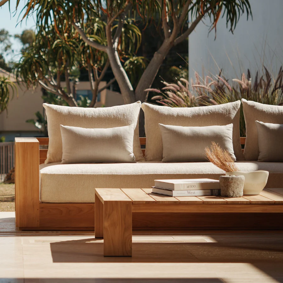 Outdoor seating area with a beige cushioned wooden sofa and backrest cushions, a matching wooden coffee table with books, a ceramic vase with dried grass, and a white bowl, set outside near trees.