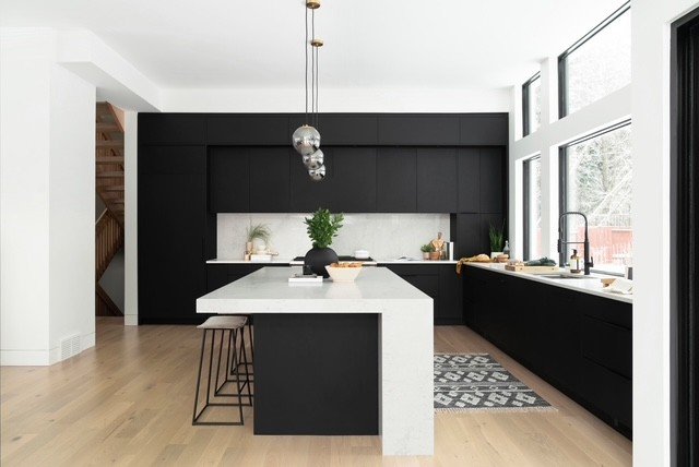 Modern kitchen with black cabinets, white marble island, pendant lights, large windows, and light wood flooring.