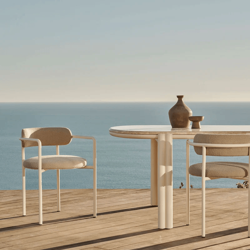 A round white table with two beige upholstered chairs on a wooden deck overlooking the ocean with a clear sky.