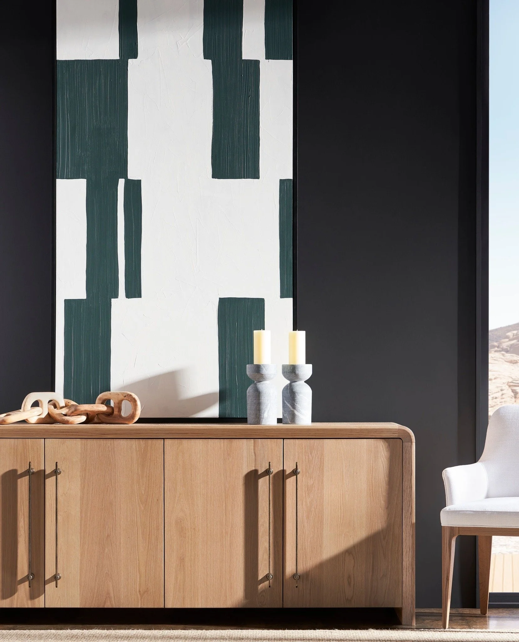 A modern living room with a wooden sideboard, decorative objects, and wall art. The sideboard has metal handles and the wall artwork features geometric shapes in green, white, and black. A white chair with wooden legs is partially visible on the right.