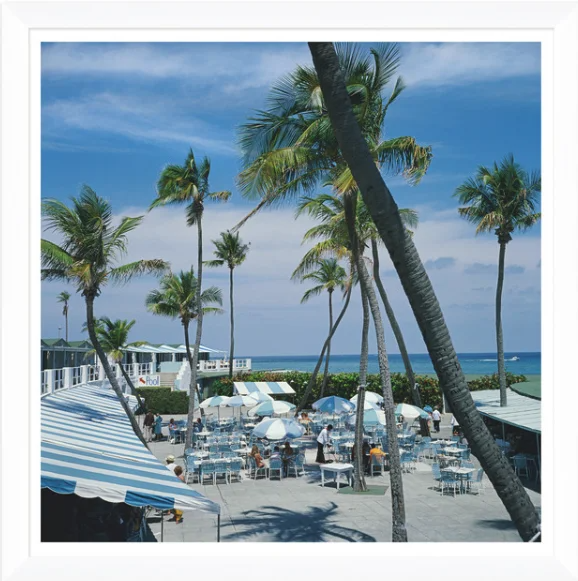 Beachside restaurant with white tables and umbrellas, tall palm trees, and a view of the ocean under a partly cloudy sky.