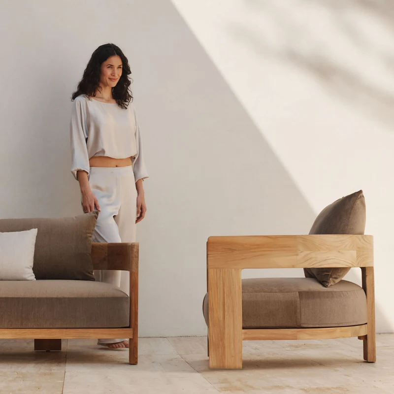 A woman standing in a minimalist living room with modern furniture, white walls, and sunlight casting shadows.