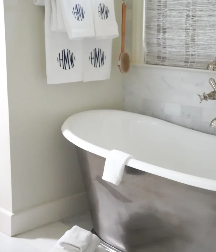A bathtub in a bathroom with a towel hanging over its edge, a rolled towel on the floor, and towels hanging on a rack with a monogrammed logo.