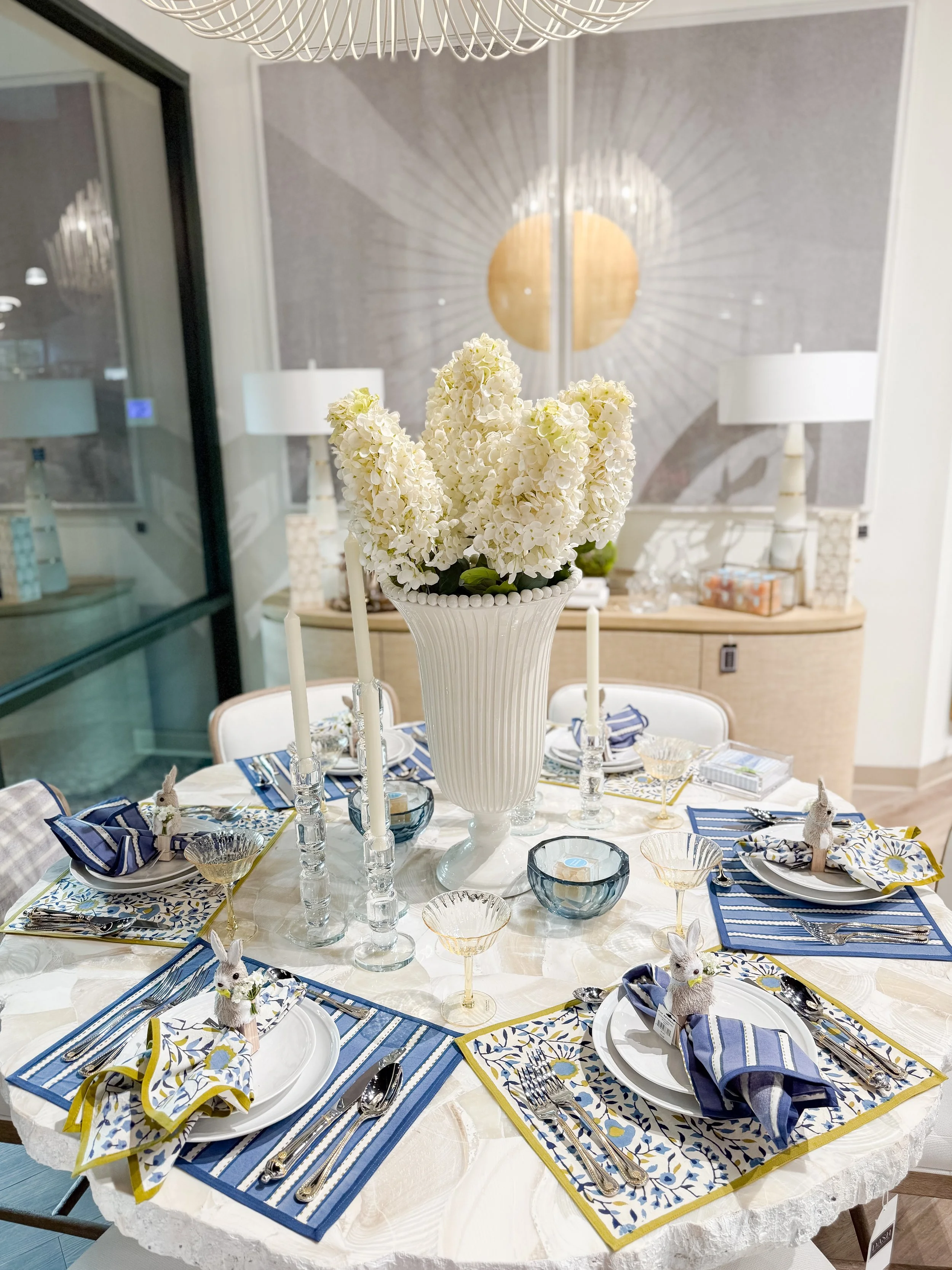A round dining table set for eight, decorated with striped and patterned navy, white, and yellow placemats, with white plates, silverware, and figurines of rabbits on each place setting. The table features a large white vase with white floral arrangements and is surrounded by glass candle holders. The background features a sideboard with lamps, ornate mirrors, and a decorative wall with a sunburst mirror.
