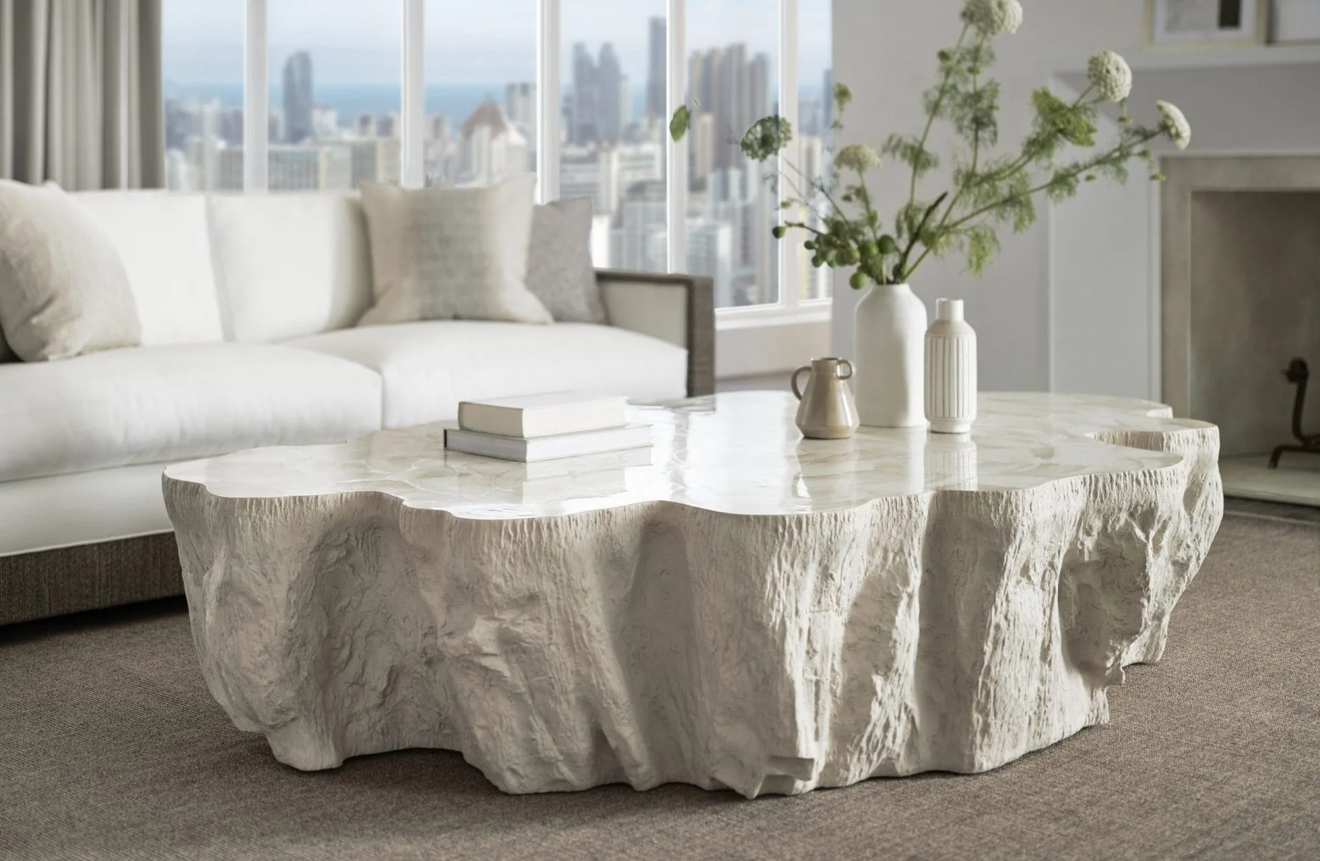 Living room with a large white sofa, a marble coffee table, and a city view through large windows. The coffee table has potted plants and books on top. The room has a modern and minimalist aesthetic.