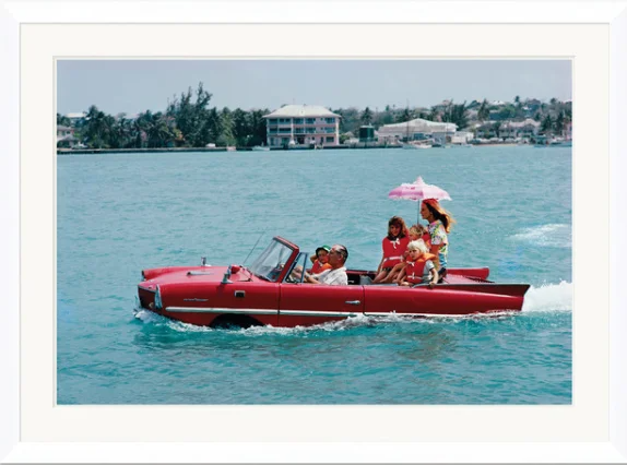 Family riding in a vintage red boat on a body of water with houses in the background, a woman holding a pink parasol.