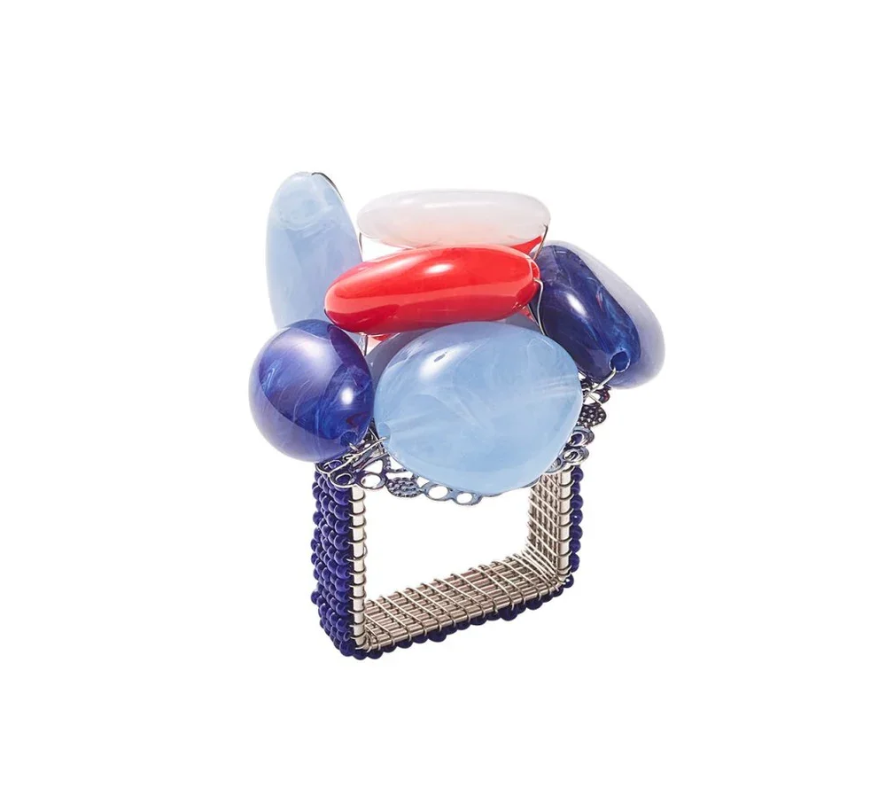 A jewelry stand with large, colorful stones including white, red, light blue, and dark blue, held in a metal wireframe cube