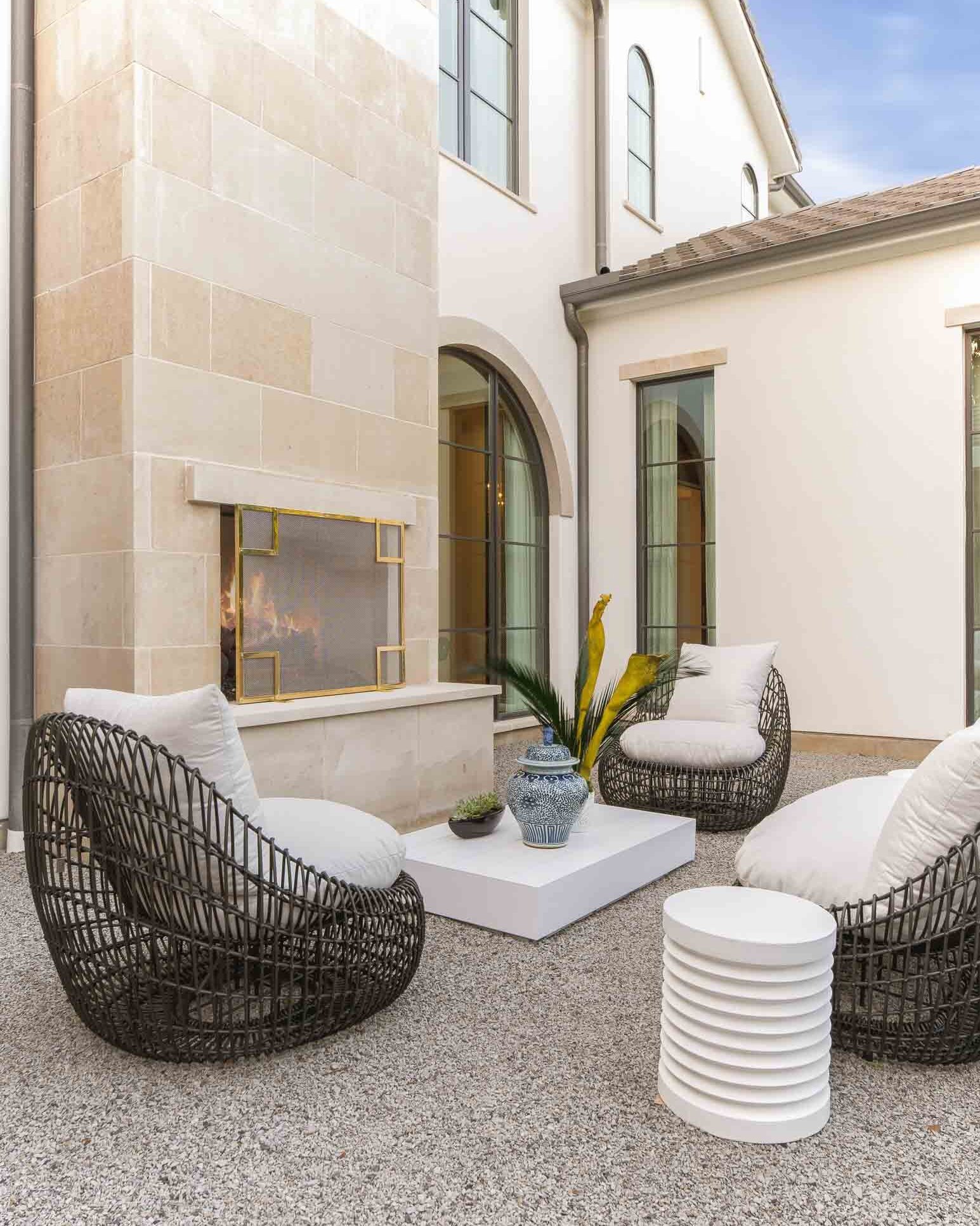 Outdoor seating area with four dark wicker chairs with white cushions, a white coffee table with blue vases, a modern white side table, and a small round white table on a gravel surface. There is a fireplace with a stone surround, a large arched wind