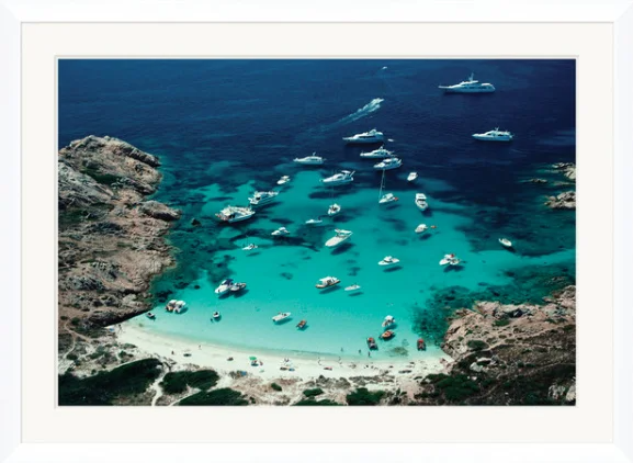 Aerial view of a cove with turquoise water, surrounded by rocky cliffs, filled with numerous boats and yachts anchored near a white sandy beach.