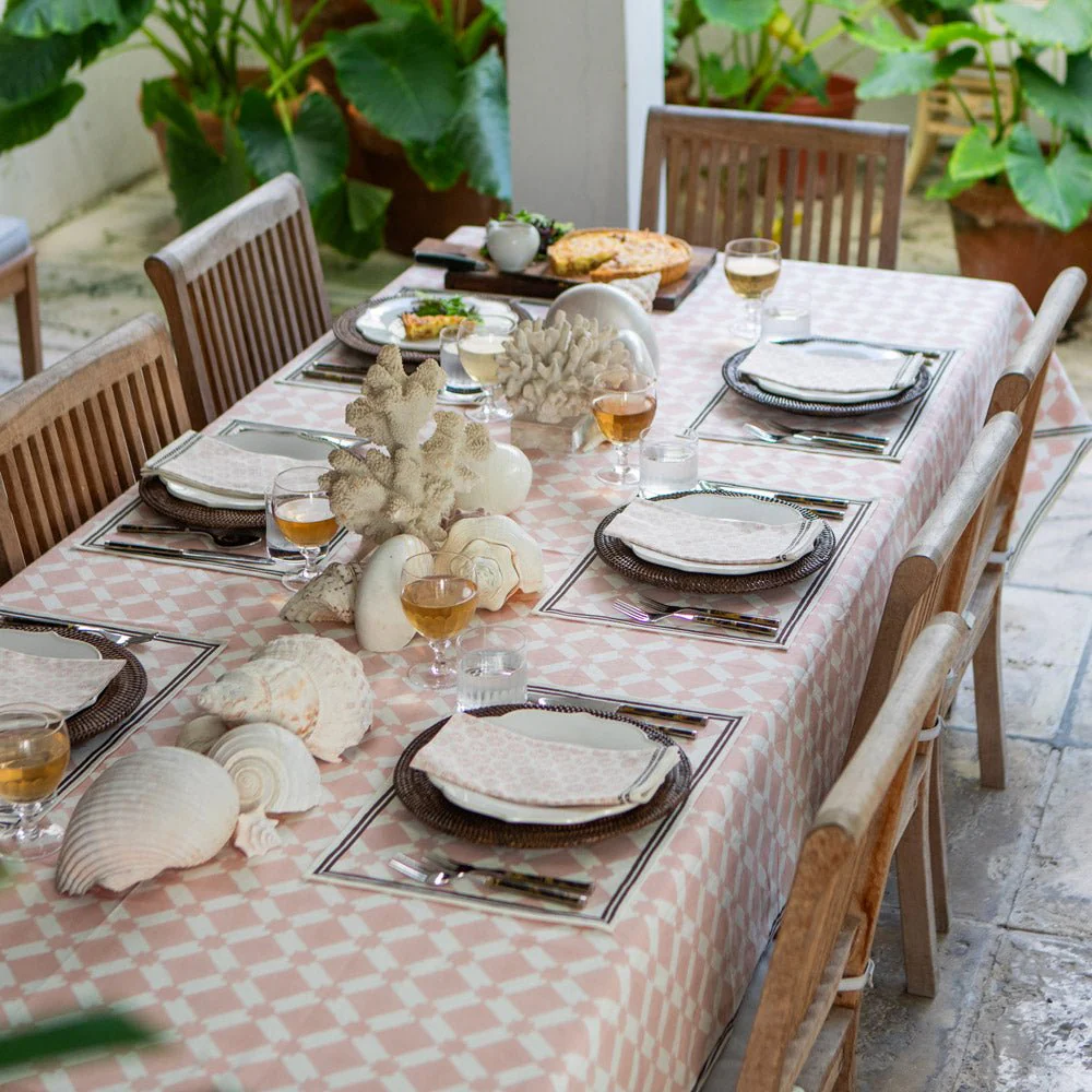 Outdoor dining table set with seashell-themed decorations, glassware, plates, and napkins, surrounded by green plants.