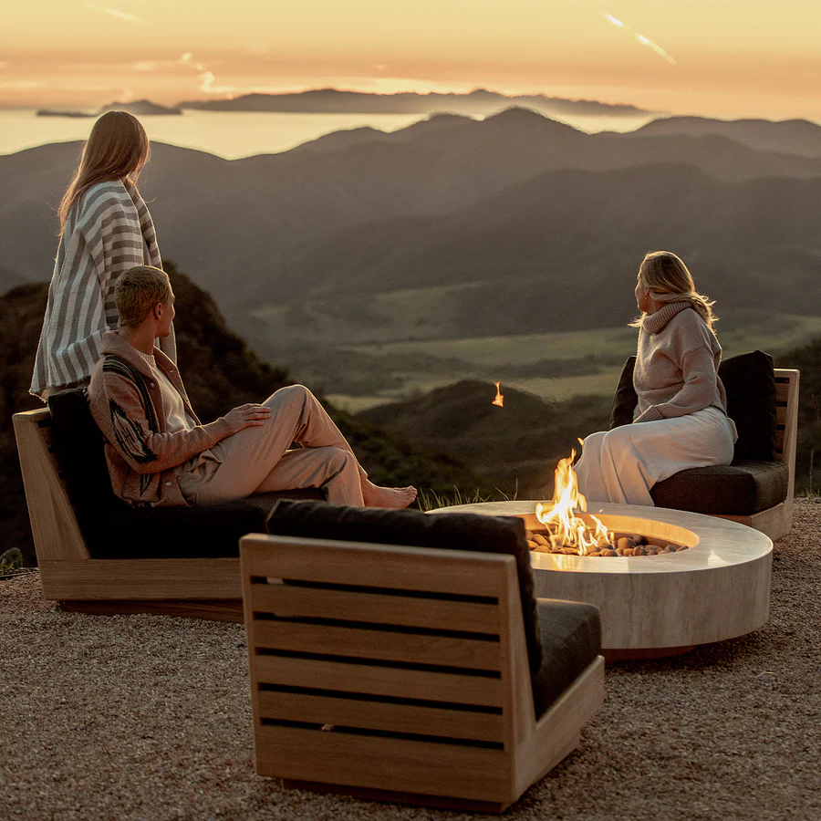 Four people gathered around a fire pit on a hillside overlooking mountains and a body of water at sunset.