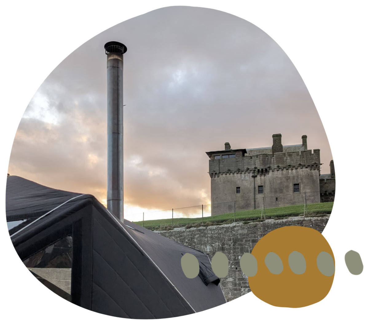 A castle with battlements on a hill with a fence in front, a tall chimney pipe, and a sauna with a black roof in the foreground. The sky is cloudy with a hint of sunlight.