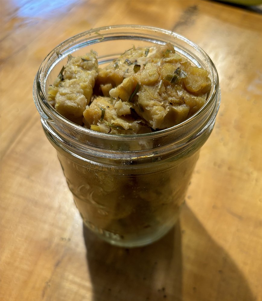 A glass jar filled with homemade chunky potato salad garnished with chopped herbs, sitting on a wooden surface.