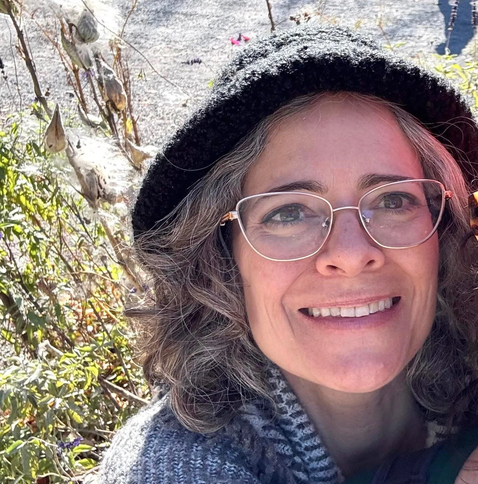 A woman smiling outdoors, wearing glasses, a black hat, and a gray sweater, with trees and plants in the background.