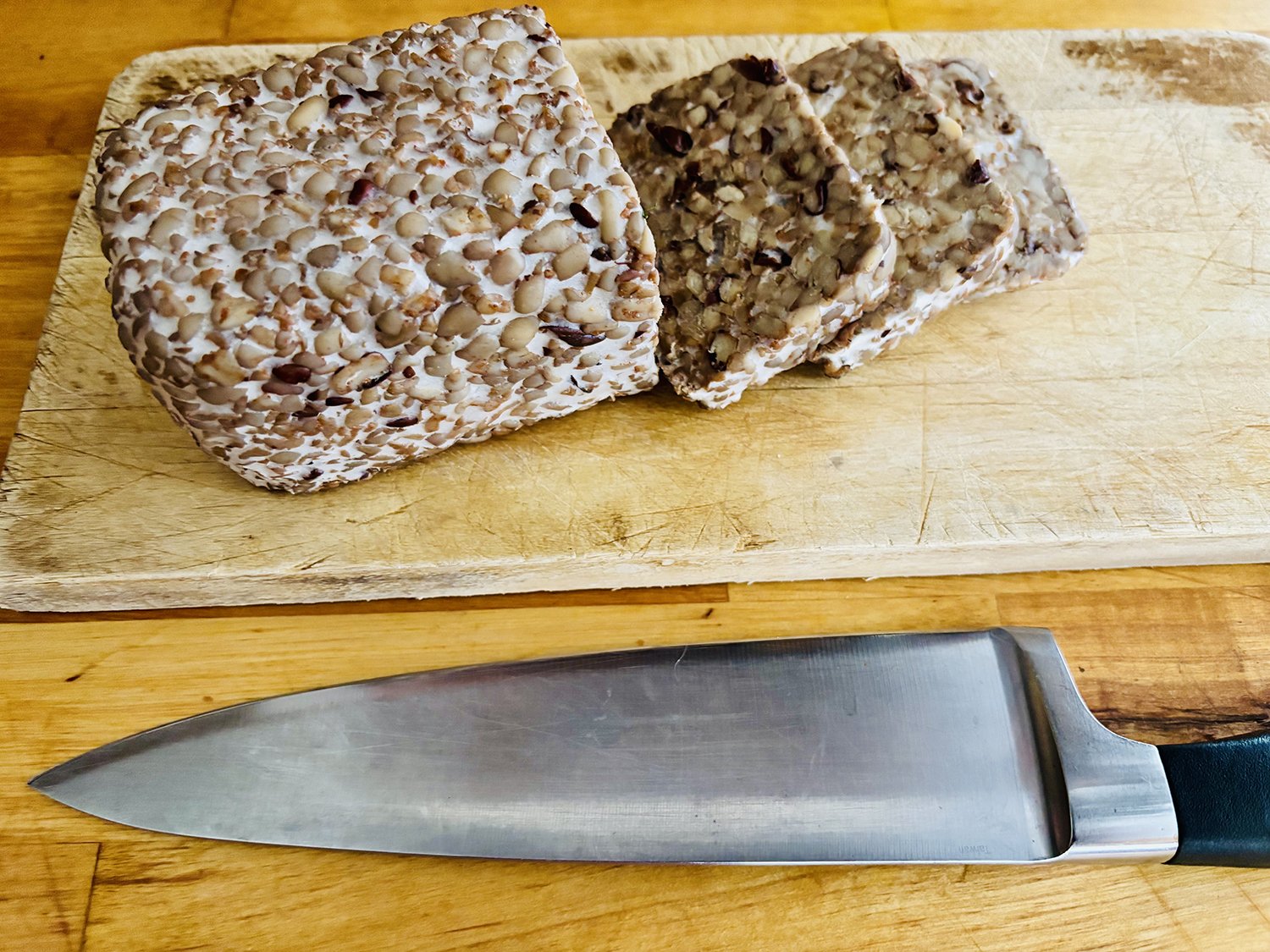 Piece of meatloaf coated with chopped nuts on a wooden cutting board with a large kitchen knife below.