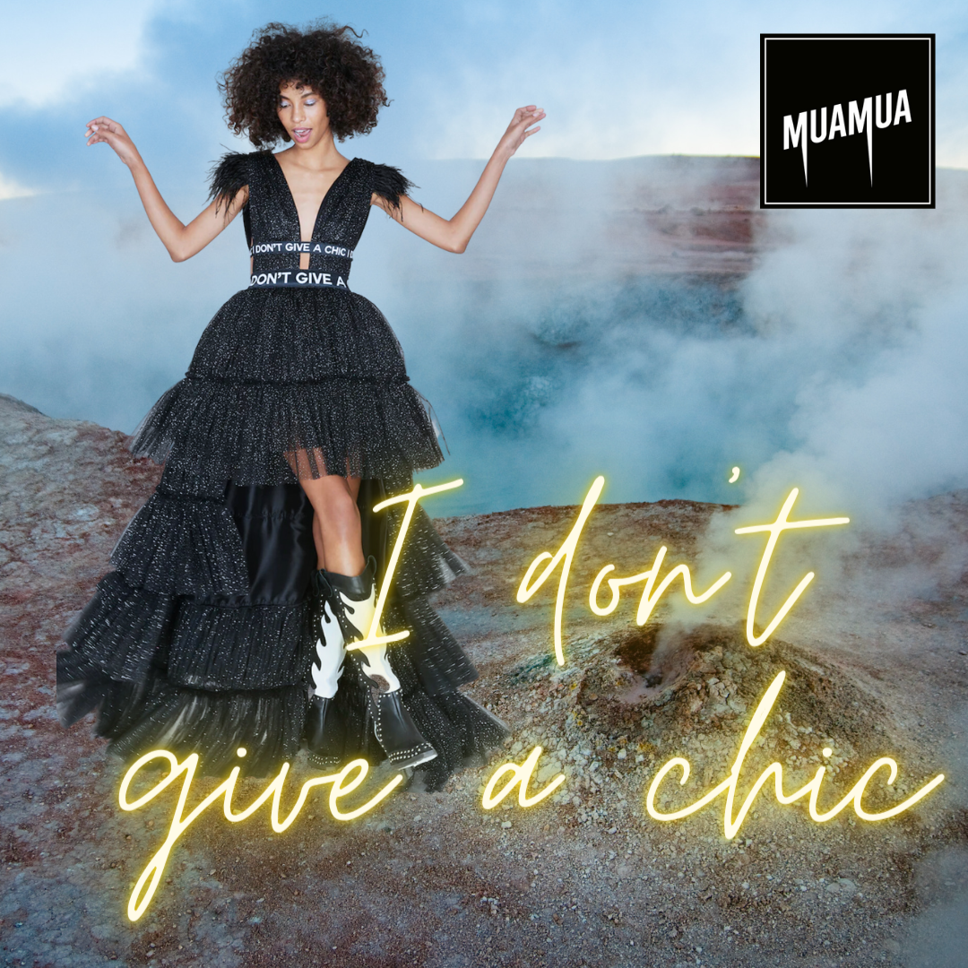 Donna che indossa un abito nero con gonna a più strati e tacchi con fiamme, in un paesaggio vulcanico con fumo, sullo sfondo. Logo di MUAMUA in alto a destra e frase luminosa 'I don't give a chic' in primo piano.