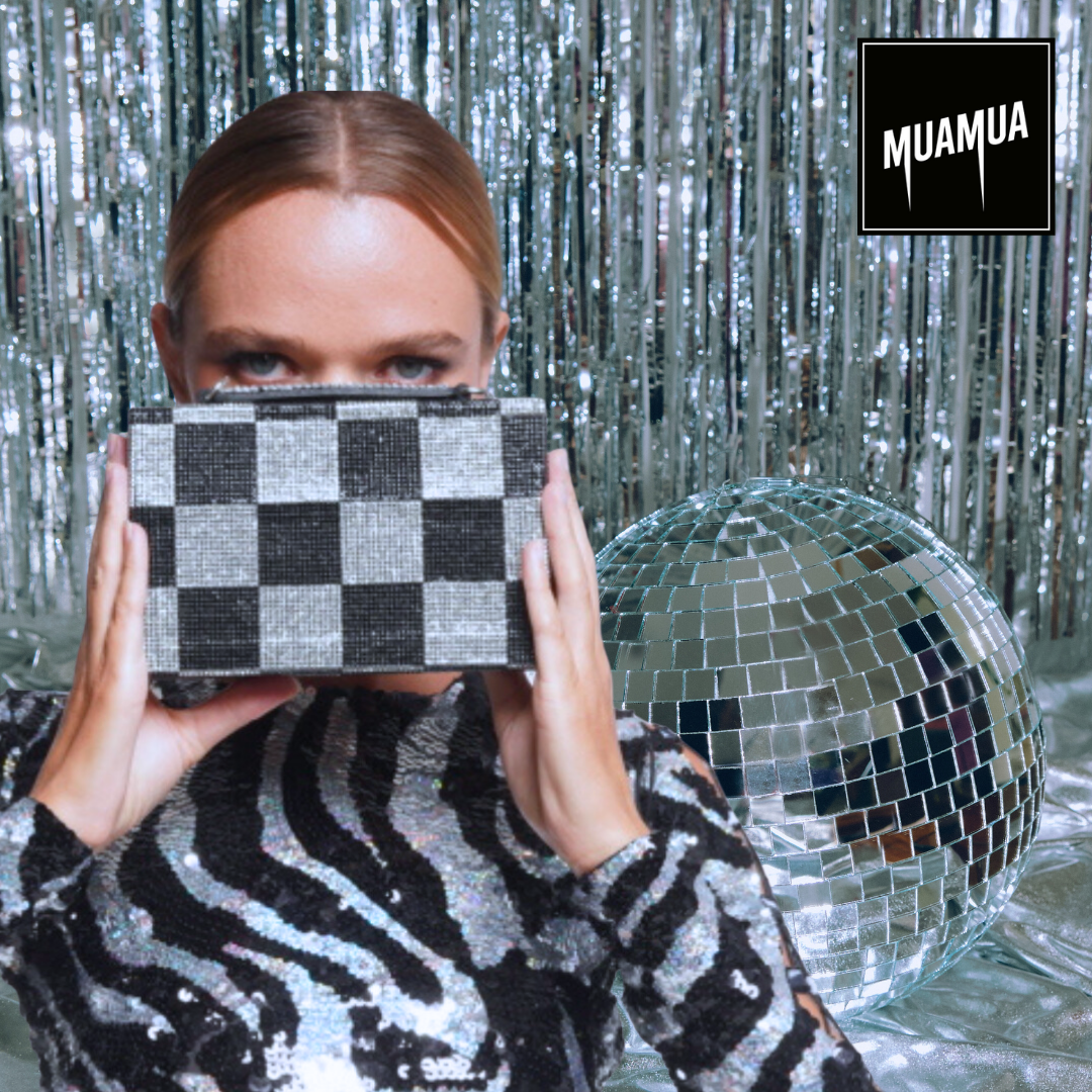 Una donna con i capelli rossi e vestito lucido con motivi zebra tiene una scatola con motivo a scacchi di fronte al volto, davanti a uno sfondo di tenda argentata e con un grande globo di specchi a forma di disco.