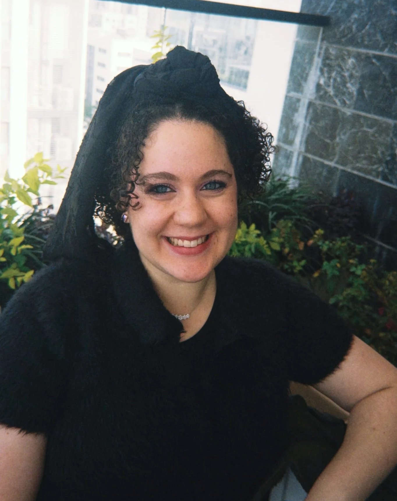 A smiling woman with curly hair and blue eyes, wearing a black top and jewelry, sitting outdoors near a glass window with plants and a cityscape in the background.