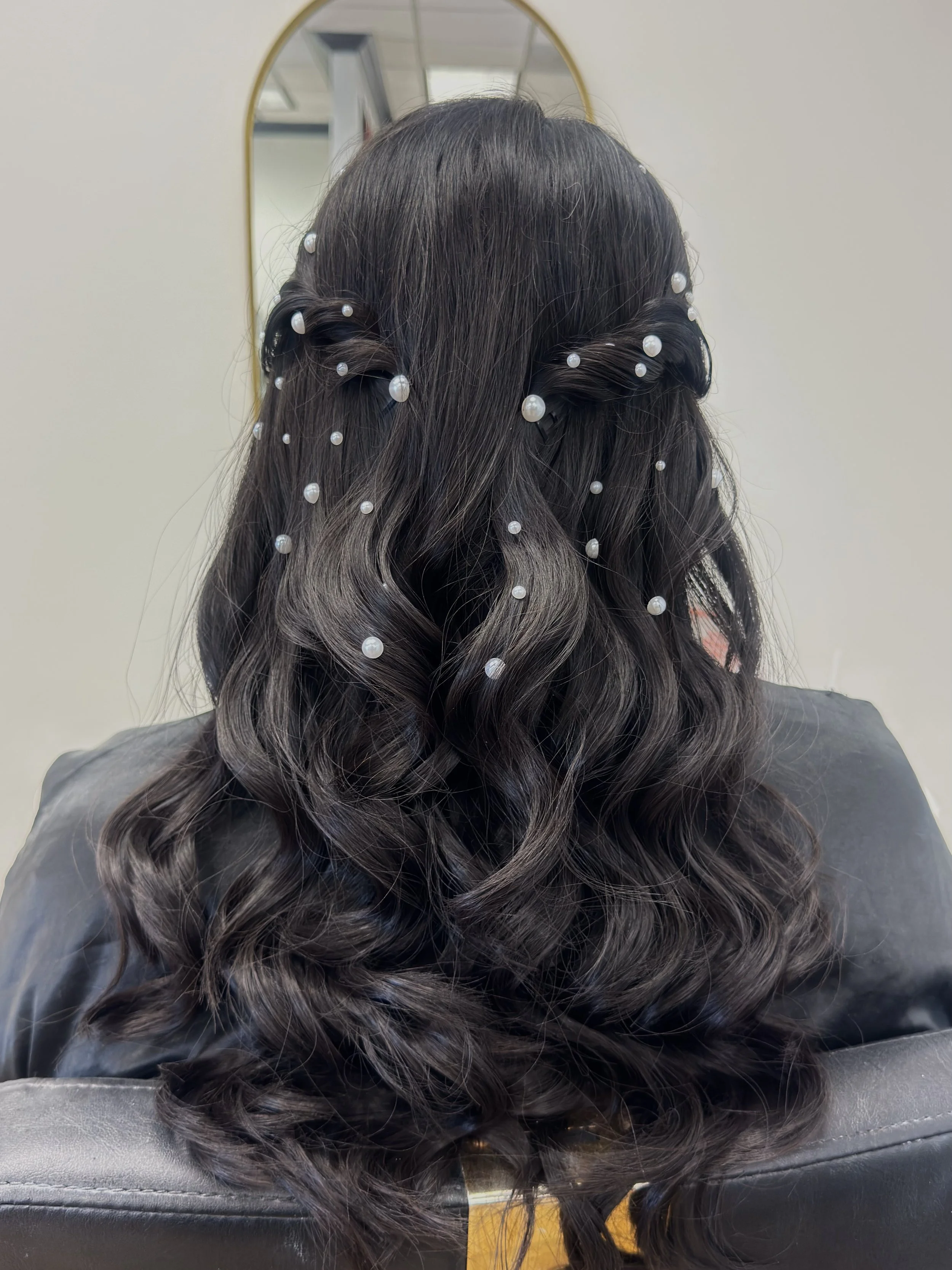 A woman with dark, wavy, and styled hair wearing a jeweled tiara. The background is dark gray.