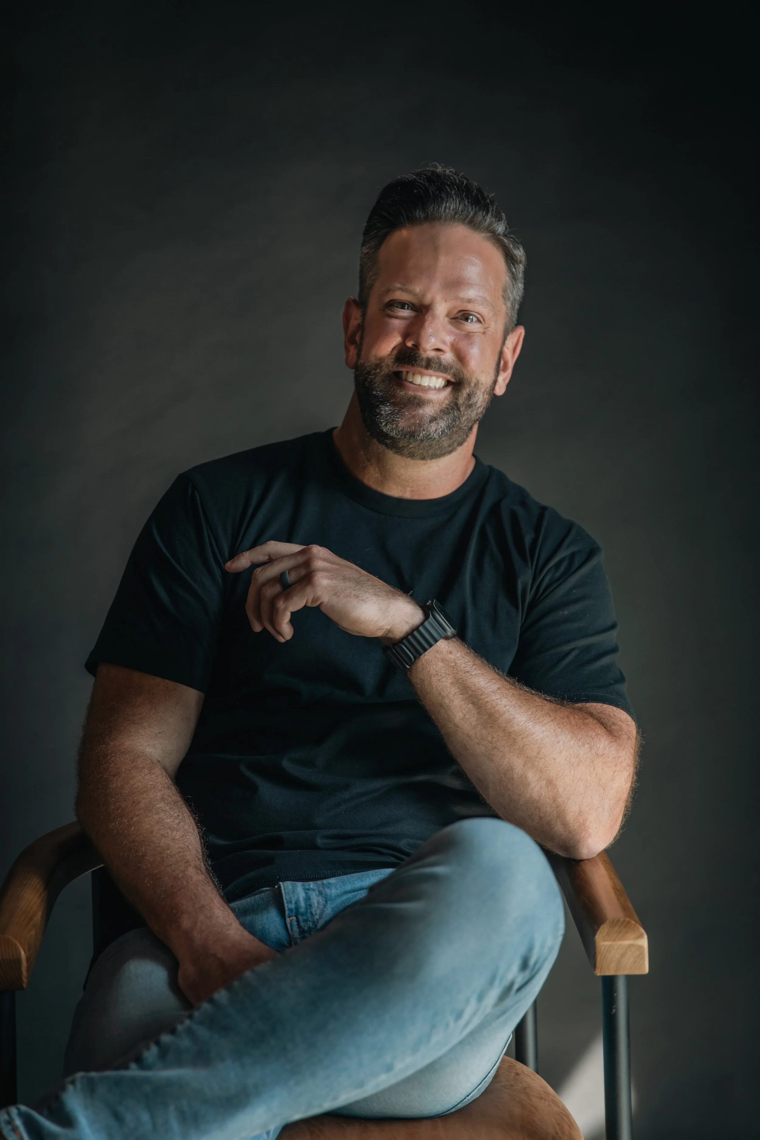 A smiling man with dark hair and beard, wearing a black t-shirt, seated in a wooden chair against a dark background.