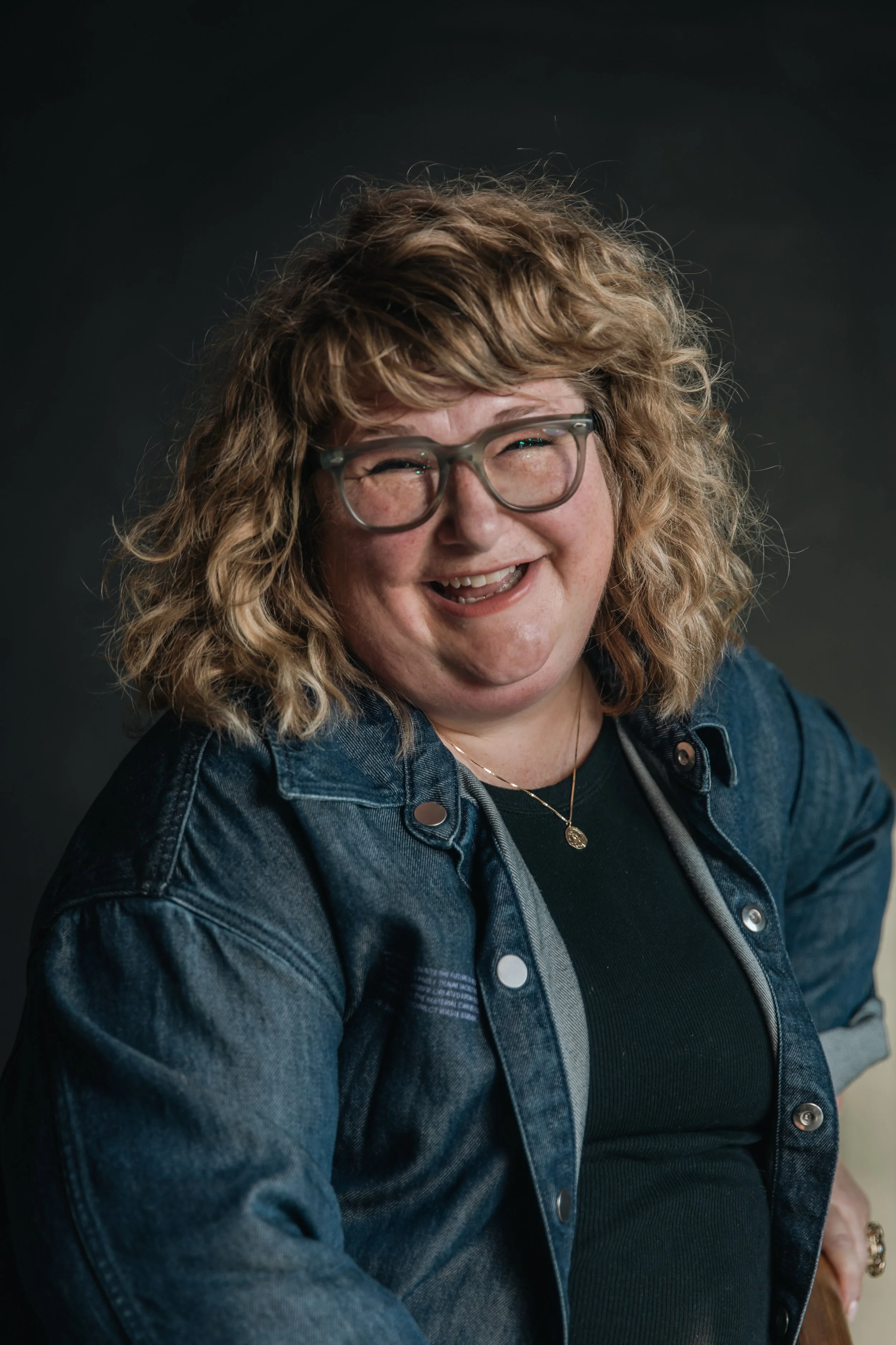 A woman with curly blonde hair, glasses, and a denim jacket smiling broadly