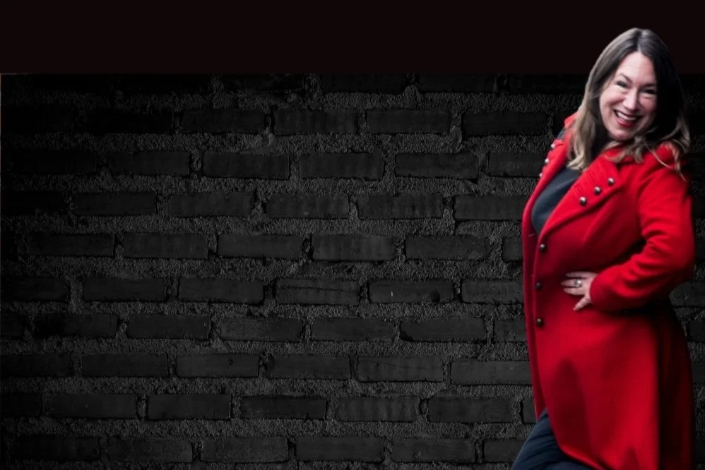 Woman in a red coat standing in front of a black brick wall, smiling energetically.