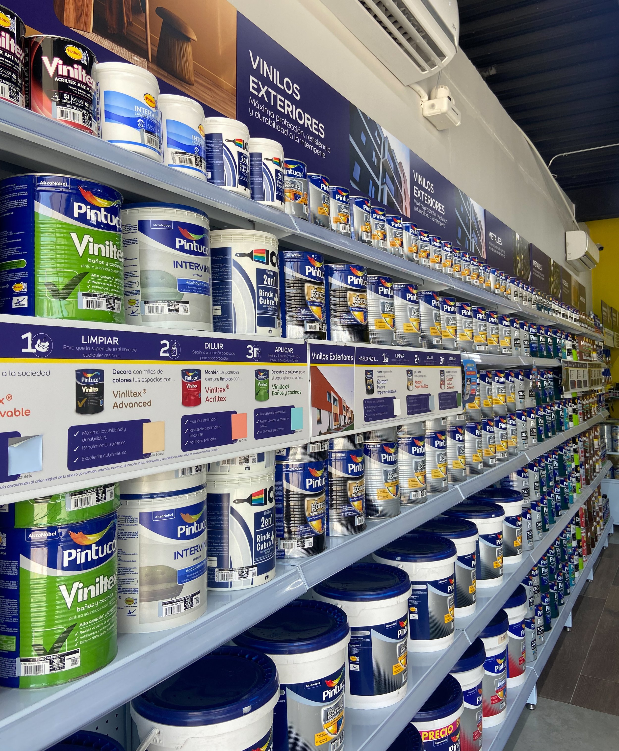 Estantería en una tienda con envases de pintura, principalmente de la marca Pintuco, en diferentes tamaños y colores, con letreros informativos en español sobre pinturas exteriores e instrucciones de uso.
