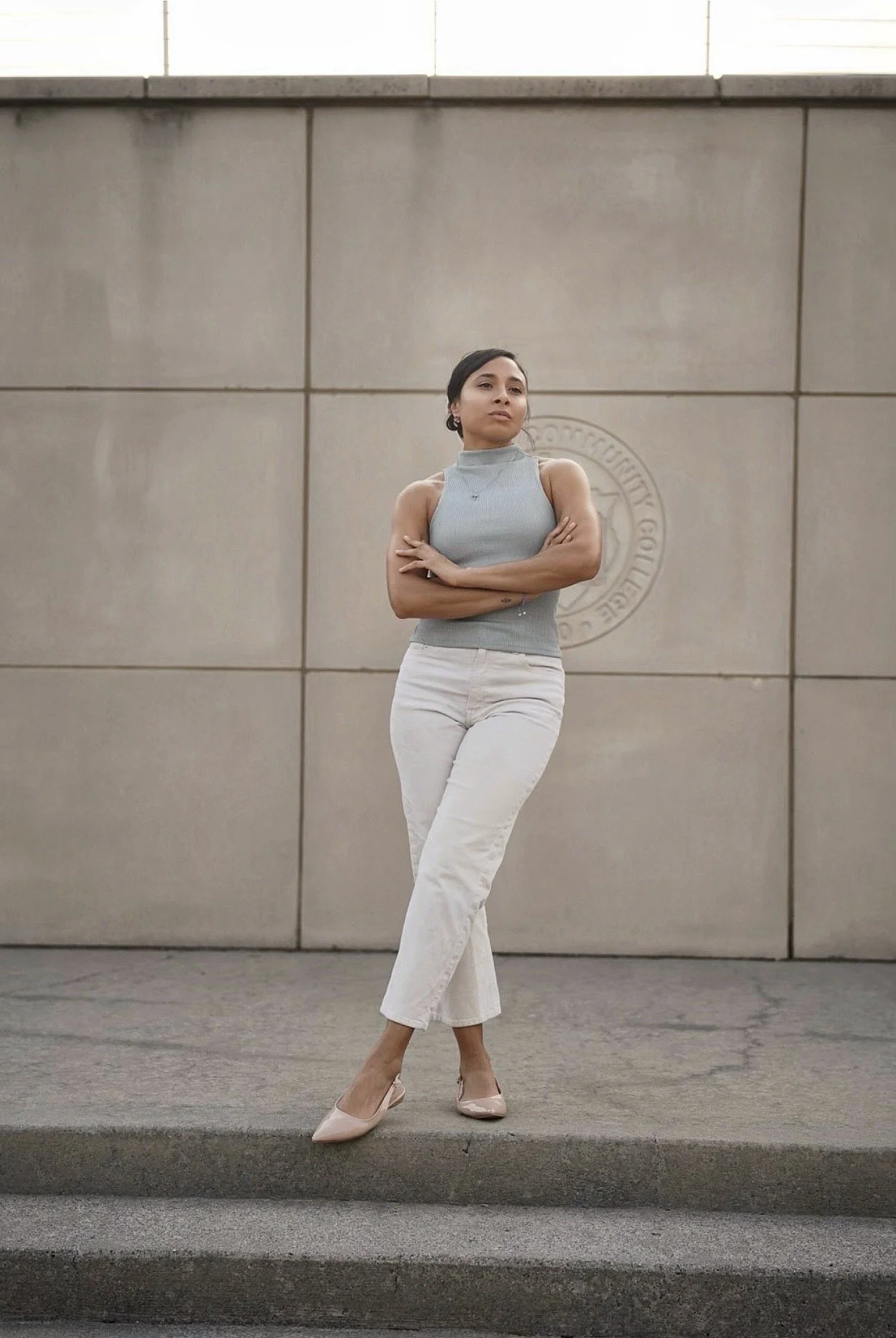 A young woman with dark hair standing against a large gray wall, dressed in a sleeveless, high-neck gray top, white pants, and nude heels, posing with her arms crossed and looking to the side.