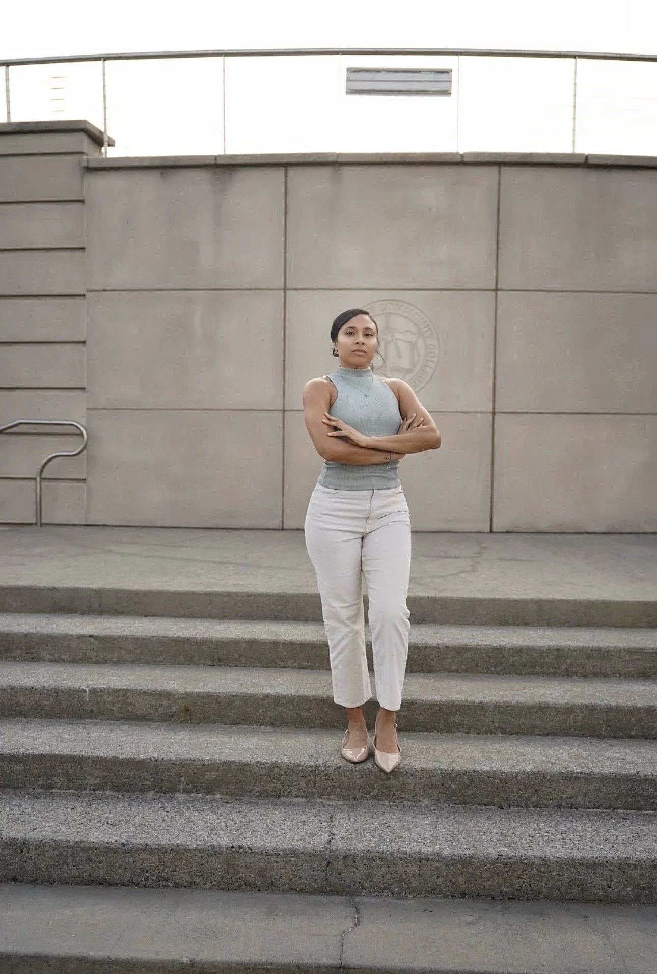 A woman in light gray sleeveless top and white pants standing on steps outside a building with a gray wall and stairs.