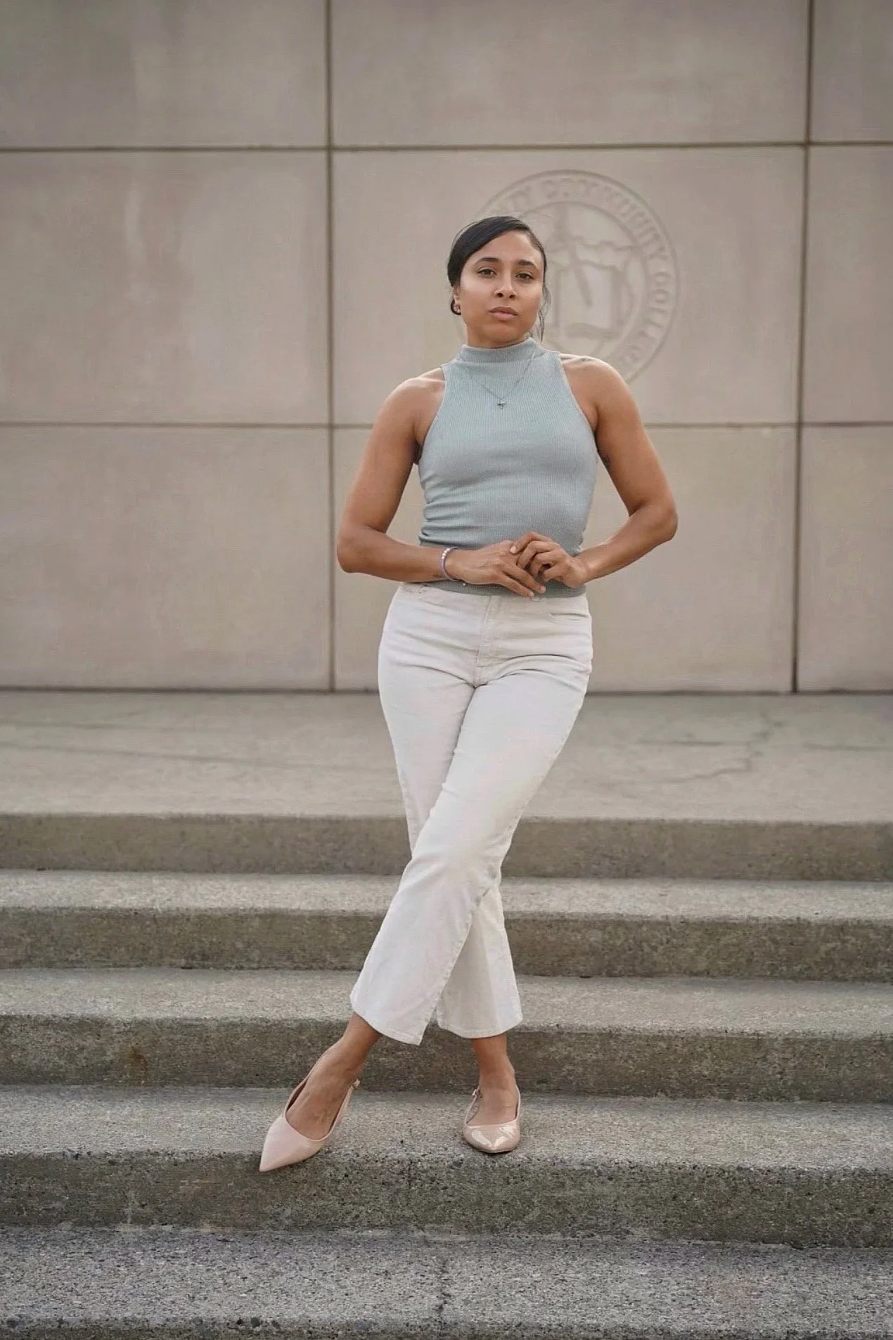 A woman standing on stairs in front of a building wall with an emblem, wearing a light gray sleeveless turtleneck top, white pants, and beige heels.