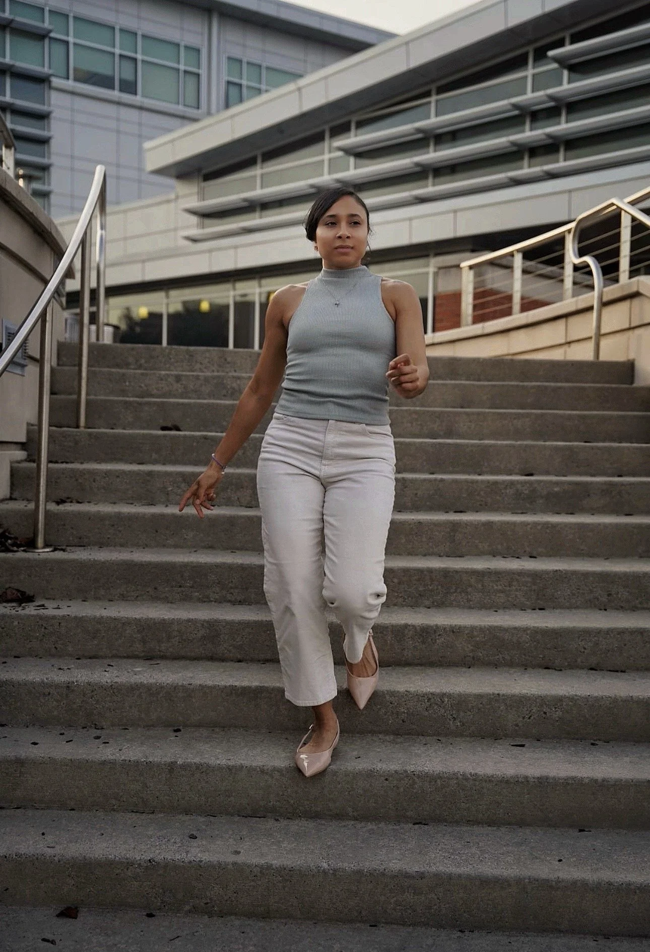 A woman walking down stairs outdoors, wearing a sleeveless gray top, white pants, and pink high heels, with modern buildings in the background.
