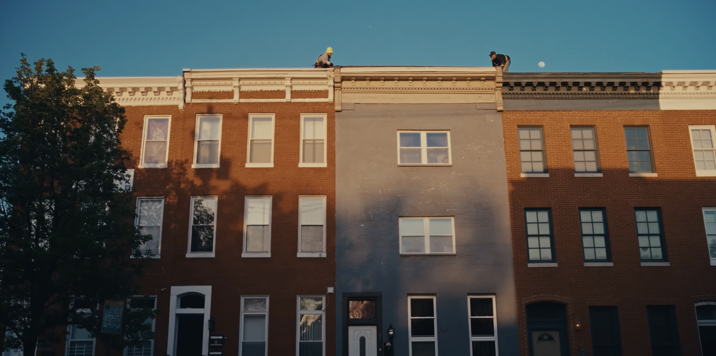 Two workers on the roof of a four-story brick building, painting the exterior wall in a gray color during early evening.
