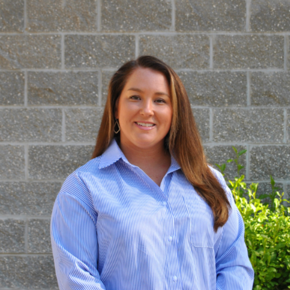 A woman with long brown hair wearing a blue and white striped button-up shirt stands outdoors in front of a grey brick wall and some green bushes.