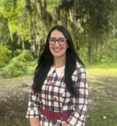 A woman with long black hair, glasses, and a checkered dress standing outdoors in a forested area.