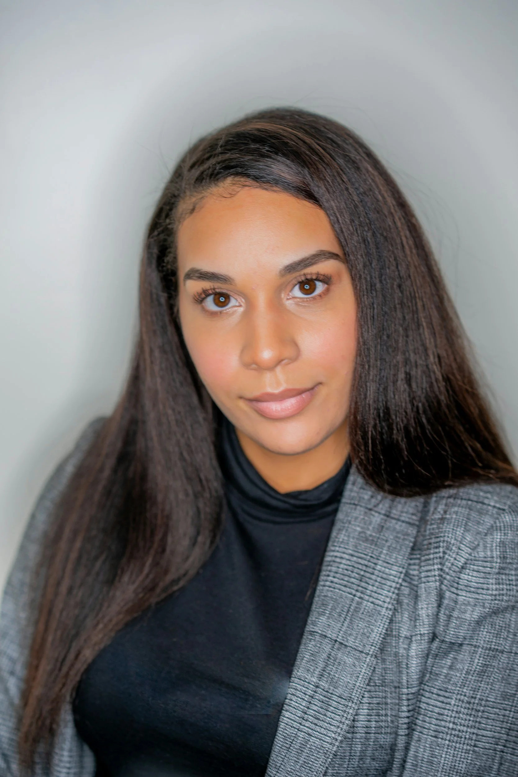 A woman with long brown hair, brown eyes, and a light complexion, wearing a black top and gray blazer, looking directly at the camera with a neutral expression.