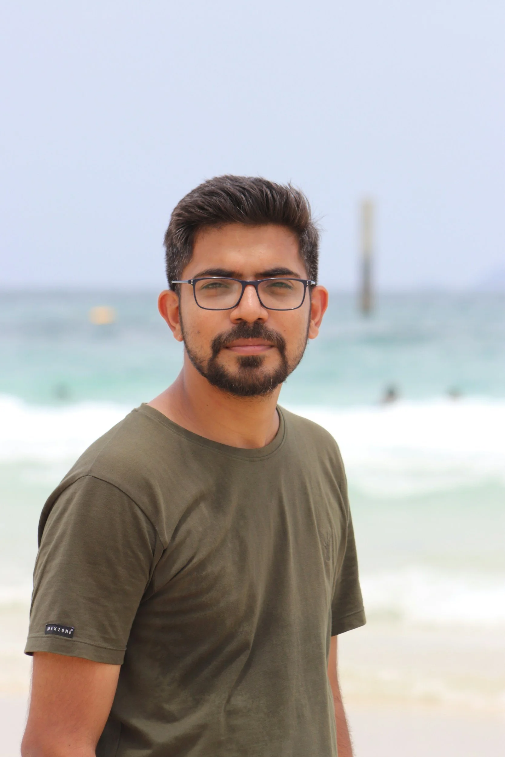A man with glasses and a beard stands on a beach, with ocean water and a buoy in the background.