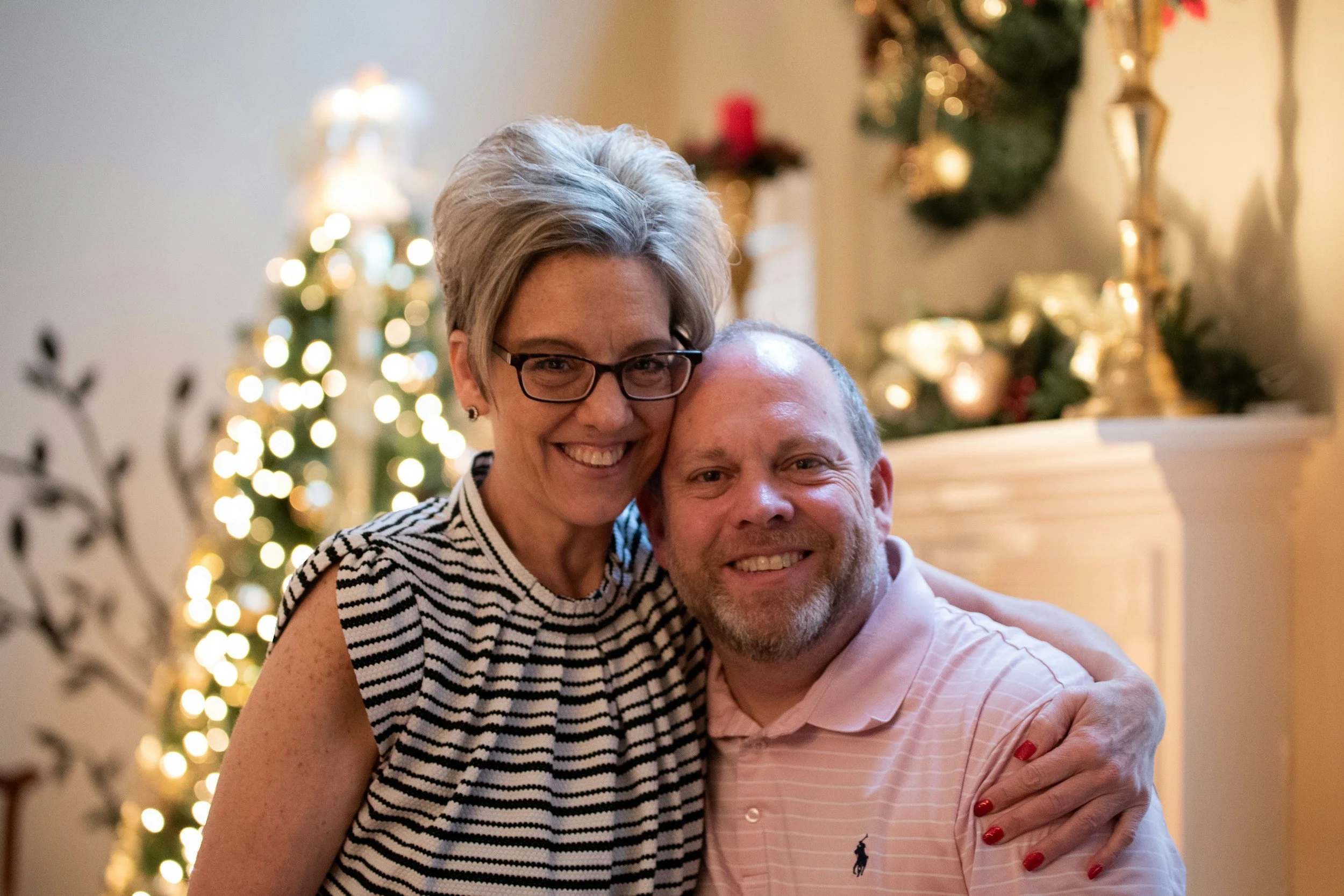 A smiling woman with short gray hair, glasses, and a sleeveless striped top hugging a smiling man with a beard and a pink polo shirt, in front of a decorated Christmas tree with lights and ornaments.