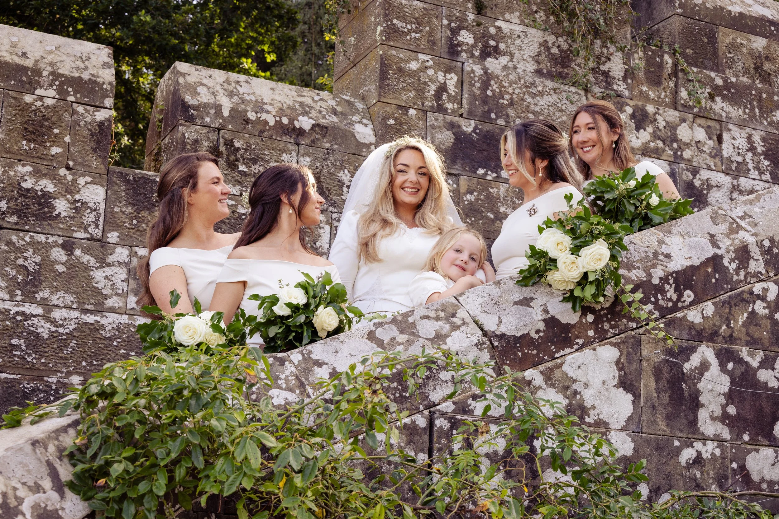 Group of women and a young girl gathered around a stone structure, smiling and holding bouquets of white roses, outdoors with greenery and moss on the stone wall.