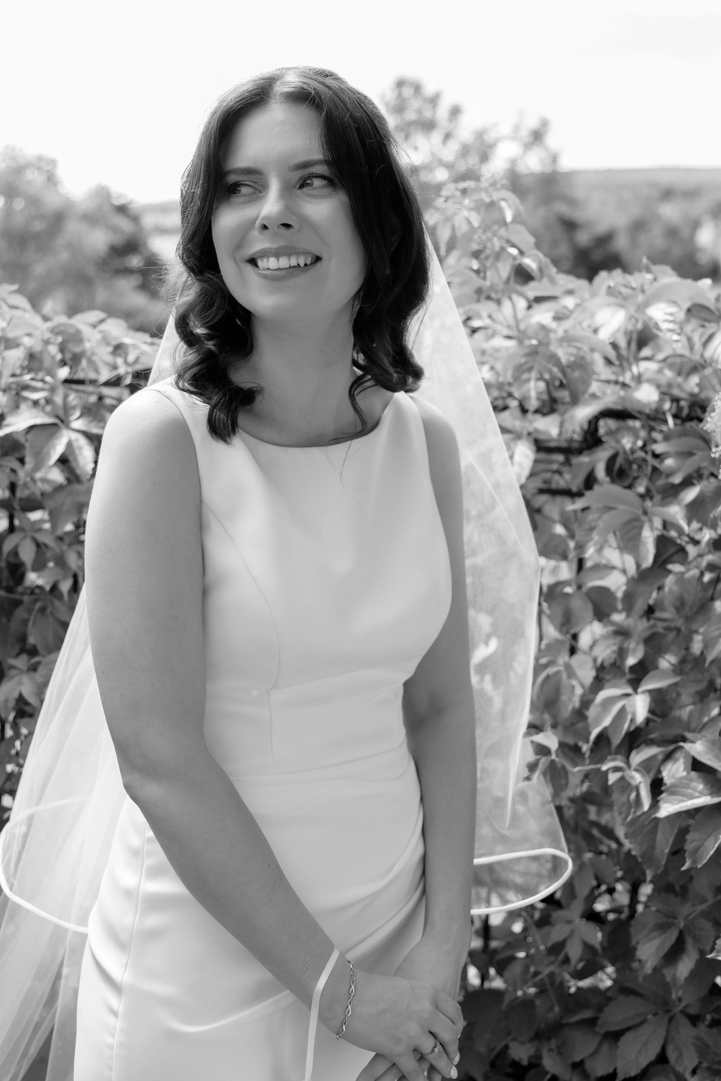 A black and white photo of a woman in a wedding dress with a veil, smiling and looking to her left, outdoors with leafy bushes and trees in the background.