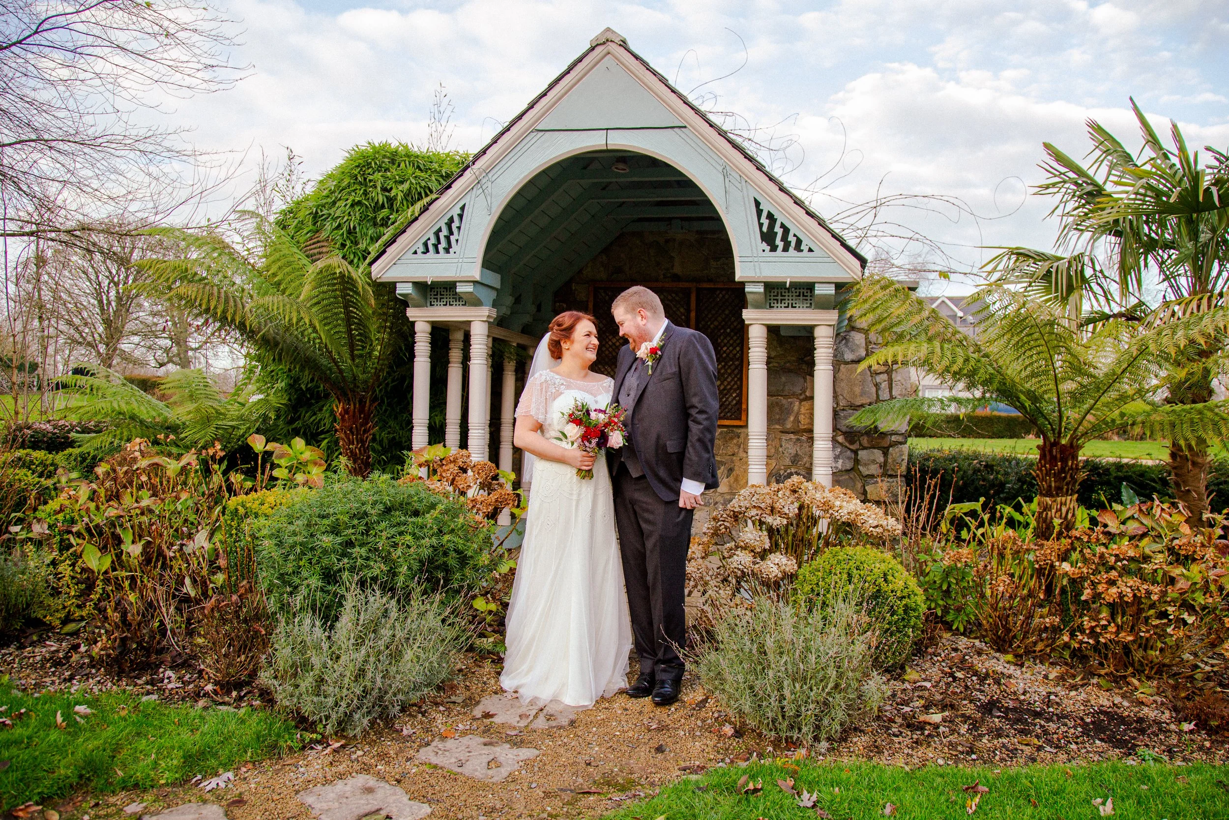 A bride and groom standing close together and smiling in a garden with lush greenery and a small wooden pavilion in the background.