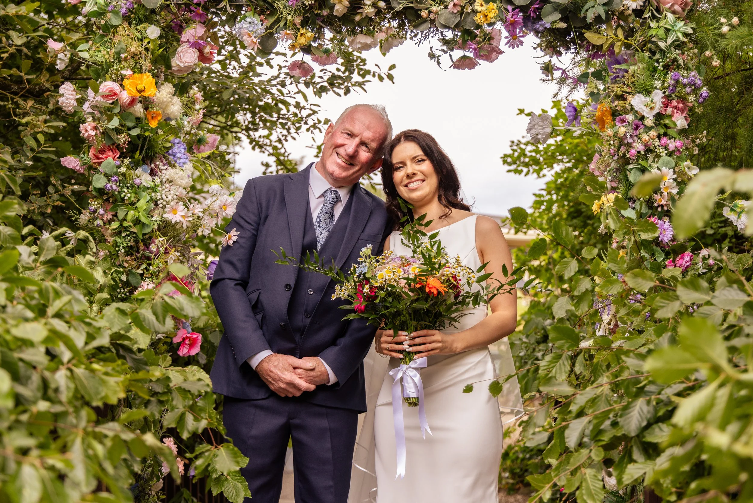 A smiling young woman in a white dress holding a bouquet of flowers, standing next to an older man in a suit, both framed by a floral archway outdoors.