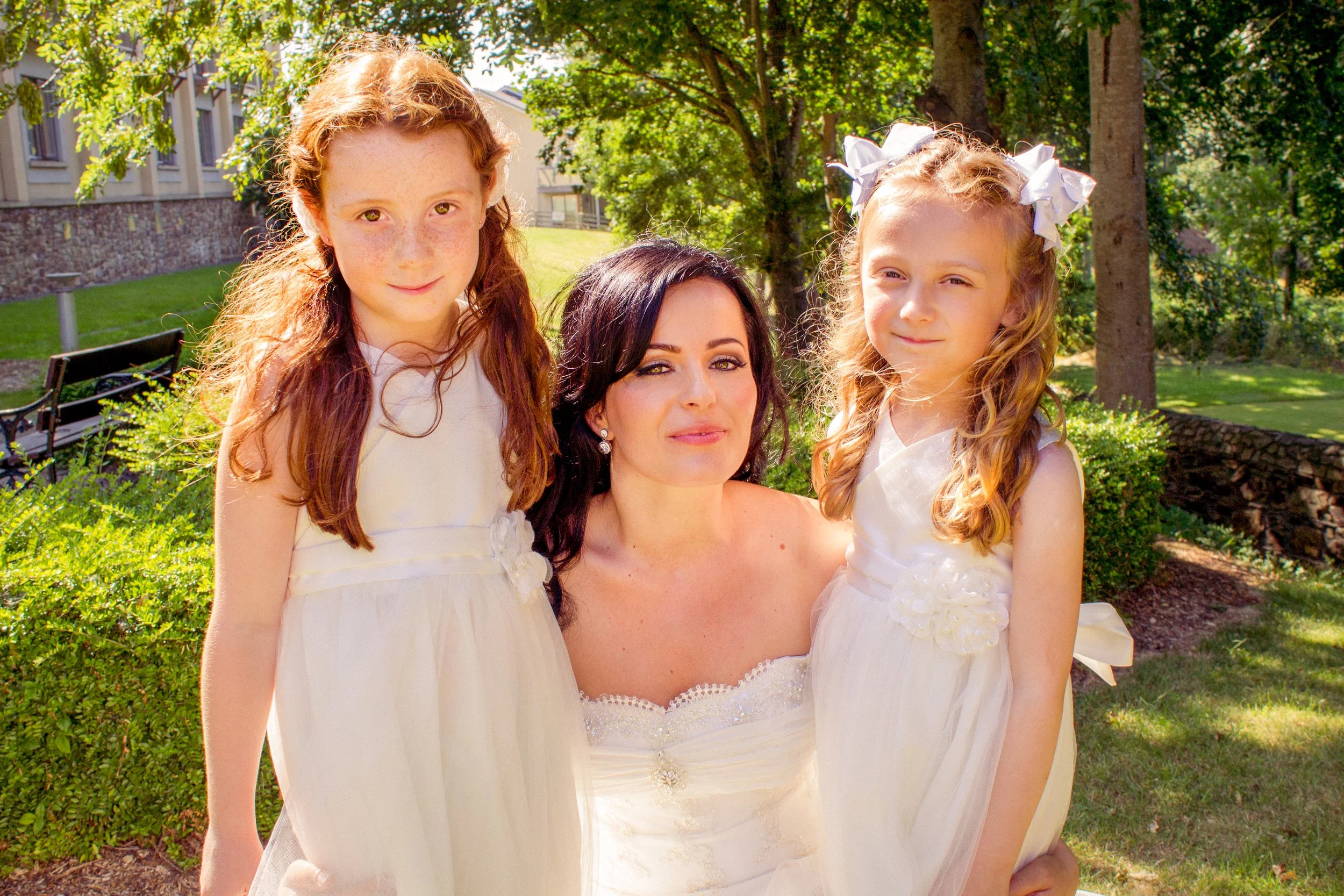 A woman with dark hair and earrings standing outdoors, flanked by two young girls in white dresses with bows in their hair, on a sunny day with green trees and grass in the background.