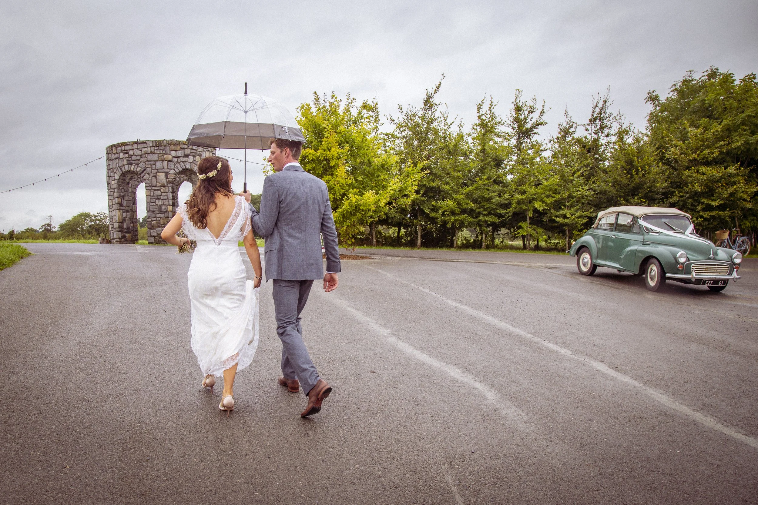 A bride and groom walking together on a paved road under an overcast sky, sharing an umbrella. The bride is in a white wedding dress with a floral headpiece, and the groom is in a gray suit. There are green trees, a vintage car, and a stone archway i