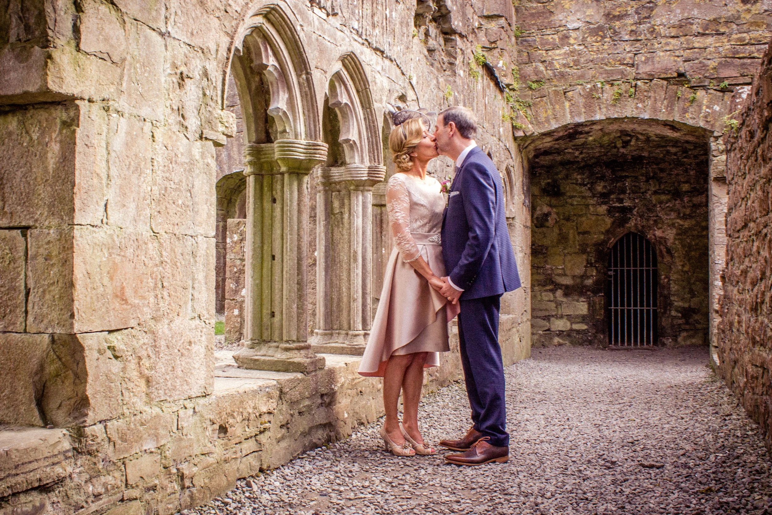 A couple dressed in formal wedding attire sharing a kiss inside an ancient stone structure with arched windows and a small barred doorway.
