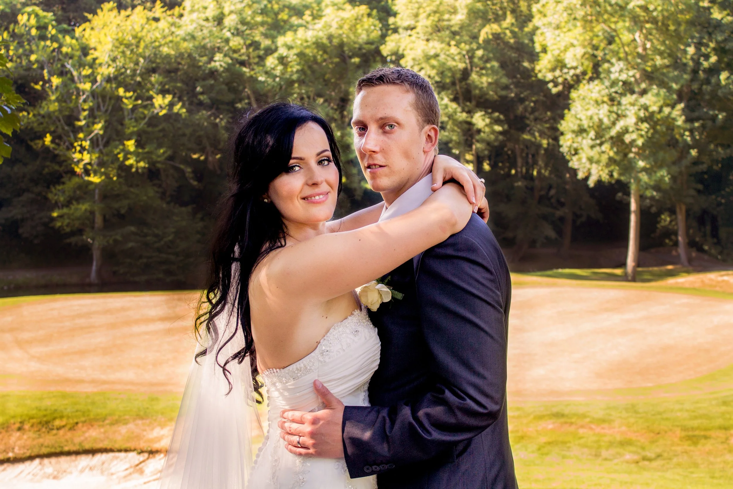 A newlywed couple standing outdoors, embracing each other, with trees and sunlight in the background.