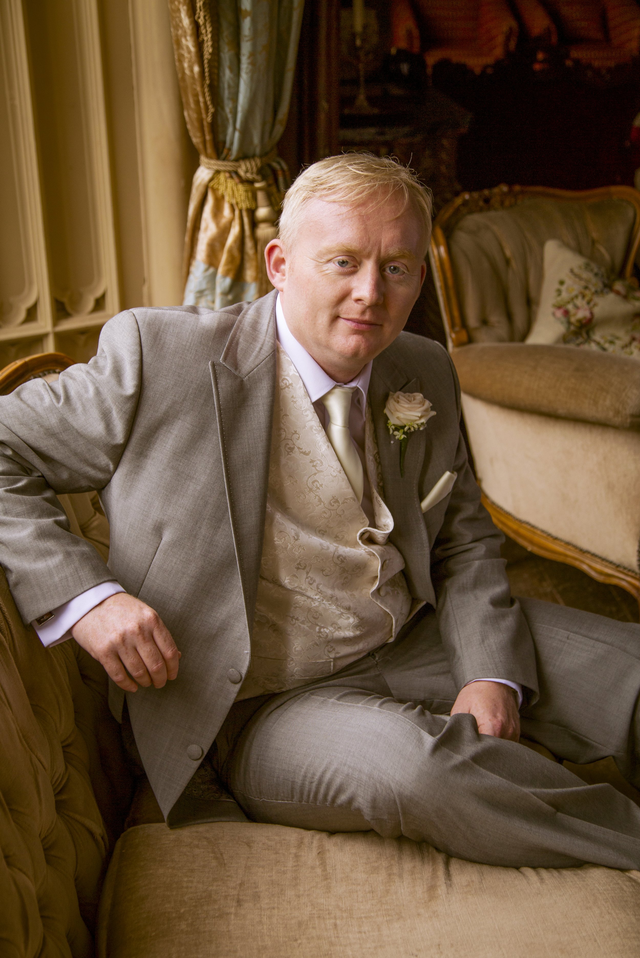 A man with blonde hair in a gray suit, white vest, and cream tie sitting on a beige vintage-style sofa in a richly decorated room with ornate furniture and gold curtains.