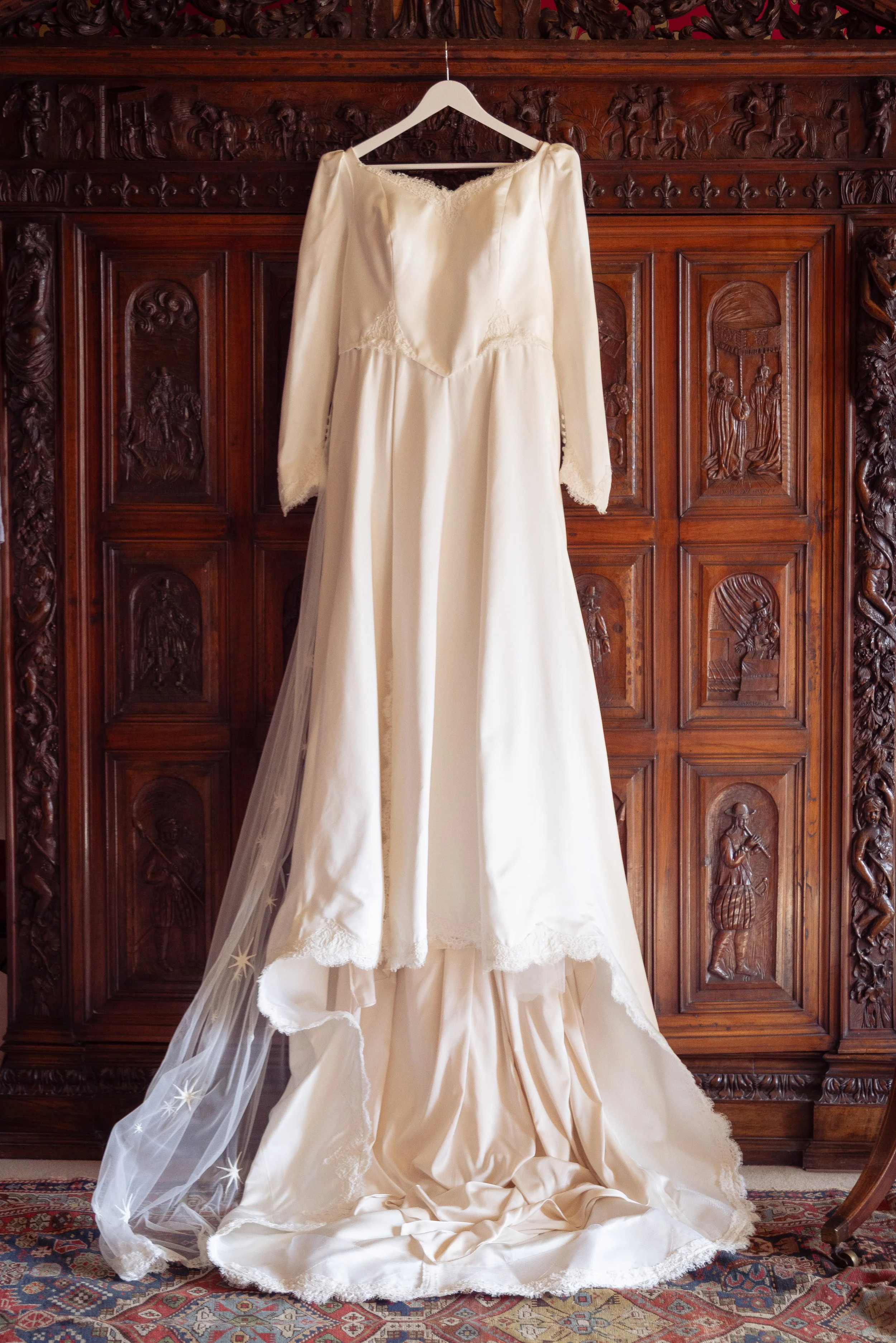 A vintage white wedding dress hanging on a hanger in front of a carved wooden wall, with a lace-edged veil draped on the floor.