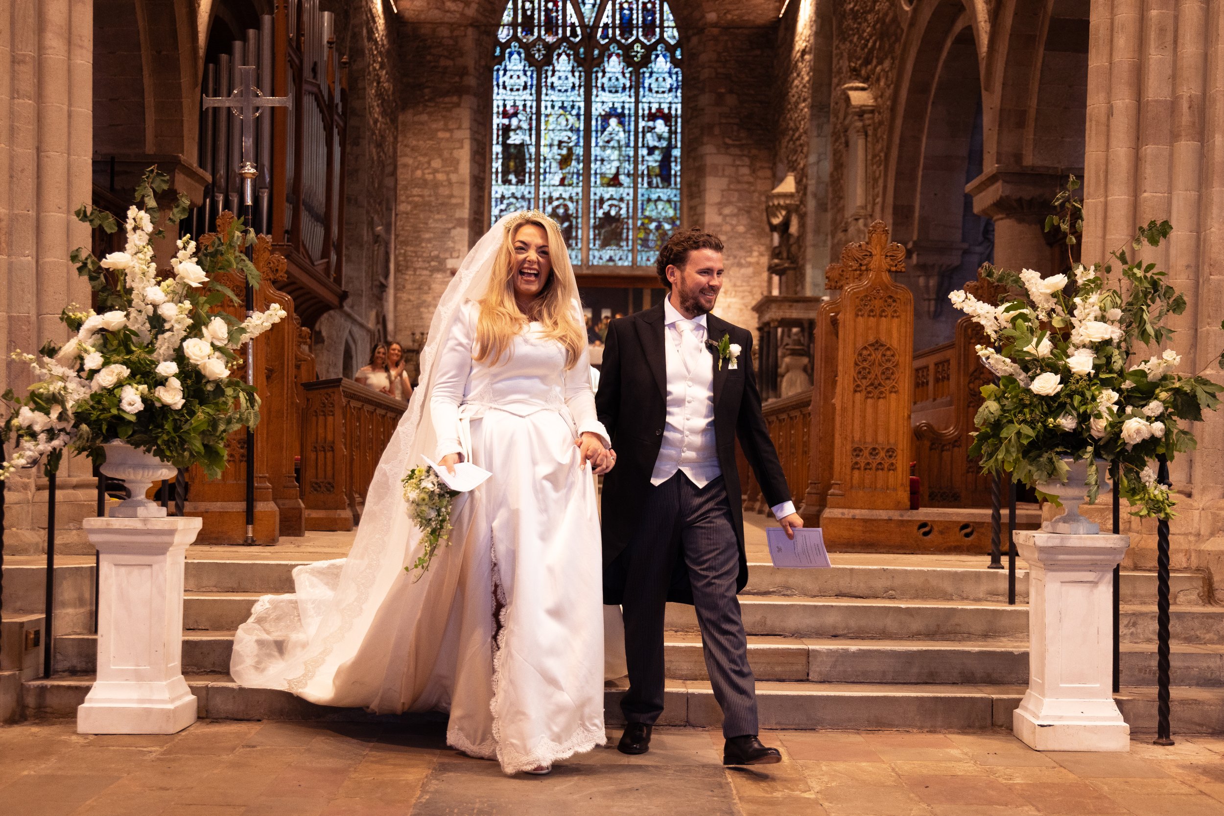 A bride and groom walking down the aisle of a church after their wedding ceremony. The bride is wearing a white wedding gown and veil, holding a bouquet, and smiling. The groom is dressed in a black tuxedo with a white shirt and boutonniere, smiling.